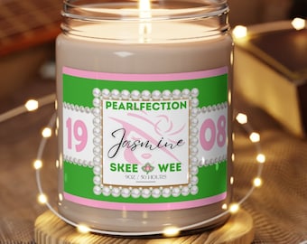 Könnte beinhalten: Eine grüne und rosa Kerze mit einem Etikett, auf dem "PEARLFECTION Jasmine SKEE WEE 9oz / 50 HOURS" steht und die Zahlen "19" und "08" auf beiden Seiten hat. Die Kerze ist angezündet und steht auf einer Holzoberfläche.