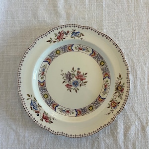 Op de afbeelding: Een ronde keramische bord met een geschulpte rand en een crèmekleurige basis. Het midden toont een bloemenboeket in roze, blauw en paars. Een decoratieve rand met ingewikkelde bloemmotieven en geometrische vormen omcirkelt de binnenste ring.