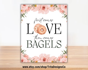 May include: A white sign with the text "first comes LOVE then comes BAGELS" in black font, with watercolor bagel illustrations. The sign is framed with pink floral accents and is displayed on a wooden surface.