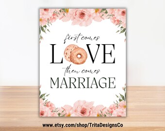 May include: A white printable sign with pink floral borders. The sign says "First comes love, then comes marriage" with a bagel and a donut in the middle of the word "love".