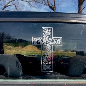 May include: A decorative cross sticker with an ornate, swirling floral design. The cross is silver and black, and is affixed to the rear window of a black vehicle. The background includes trees and a blue sky.
