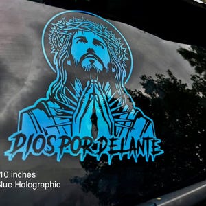 May include: Light blue holographic decal, 10 inches in size, featuring an image of Jesus with hands clasped in prayer. The text "DIOS POR DELANTE" is below the image. The decal is designed for application on a vehicle window.