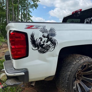 May include: A white pickup truck features a black vinyl decal on the rear quarter panel depicting two roosters in a dynamic fighting stance with wings spread. The truck has a red Z71 Off Road emblem above the decal and a large red taillight.