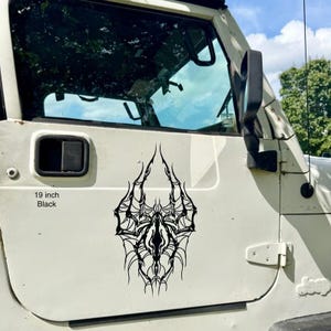 May include: A black 48 cm vinyl decal with an elaborate, symmetrical design, possibly a spiderweb or abstract tribal art. The decal is affixed to the white door of a vehicle, with the text "19 inch Black" visible.