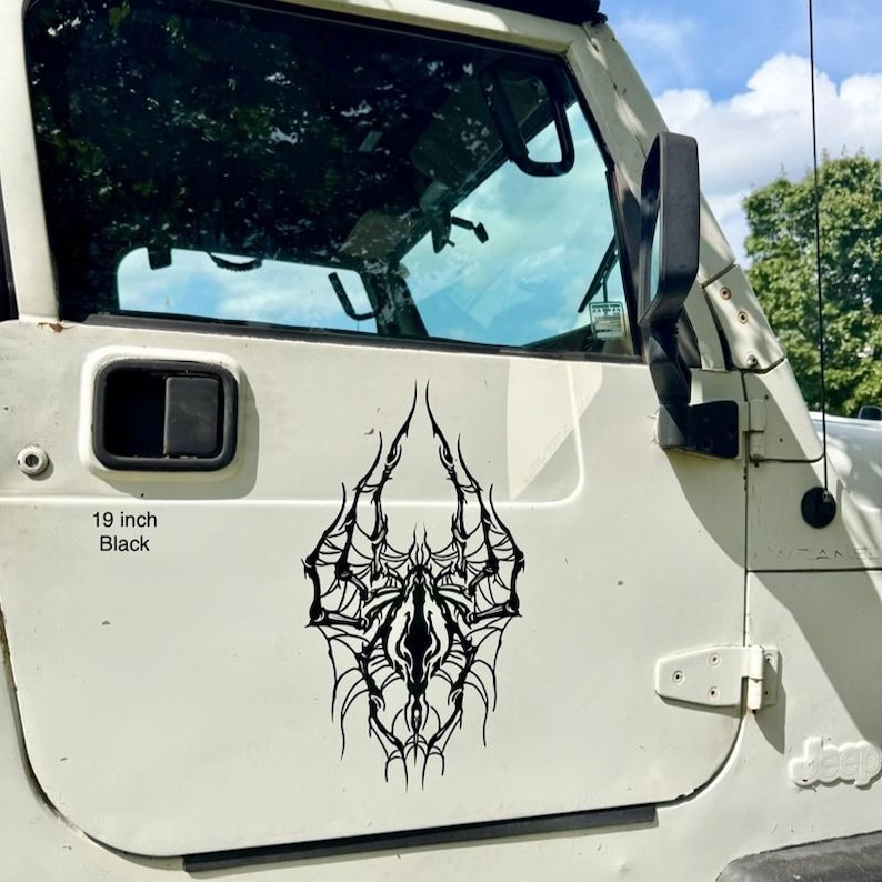 May include: A black 48 cm vinyl decal with an elaborate, symmetrical design, possibly a spiderweb or abstract tribal art. The decal is affixed to the white door of a vehicle, with the text "19 inch Black" visible.