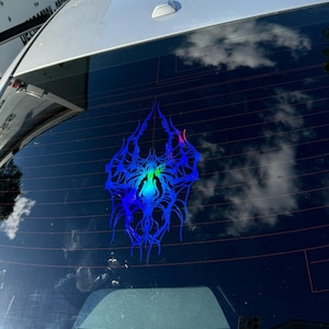 May include: A blue and iridescent car window sticker with a symmetrical, stylized design. The sticker has a central, glowing area with green and blue hues, surrounded by intricate, spiky lines. The sticker is affixed to a car window.