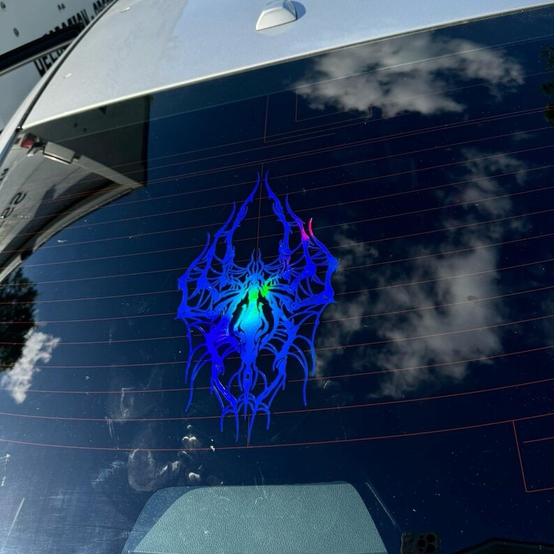 May include: A blue and iridescent car window sticker with a symmetrical, stylized design. The sticker has a central, glowing area with green and blue hues, surrounded by intricate, spiky lines. The sticker is affixed to a car window.