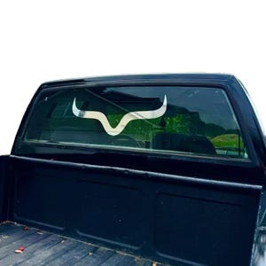 Puede incluir: Una calcomanía plateada con cuernos de toro está adherida a la ventana trasera de una camioneta negra. La calcomanía es una representación estilizada de los cuernos de un toro, con un diseño curvo. La caja de la camioneta es visible debajo.