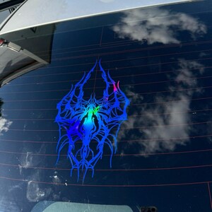 May include: A blue and iridescent spider web decal is affixed to a car window. The design features intricate, spidery lines with a gradient of colours, including blue, green, and pink. The background shows a reflection of the sky and clouds.