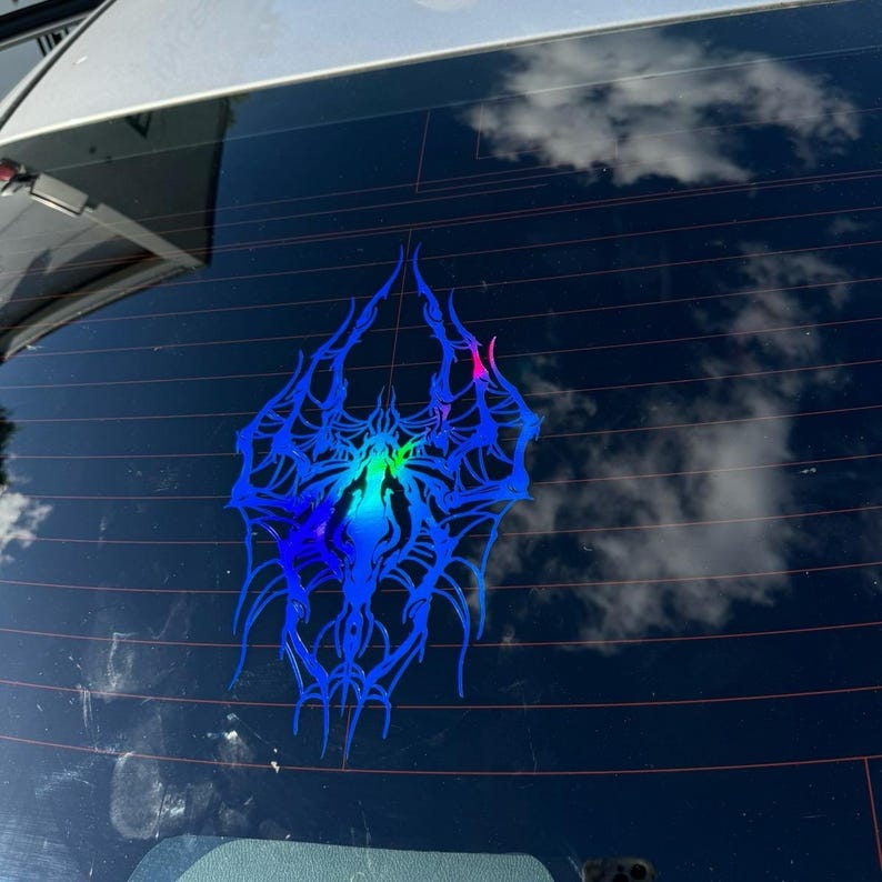 May include: A blue and iridescent spider web decal is affixed to a car window. The design features intricate, spidery lines with a gradient of colours, including blue, green, and pink. The background shows a reflection of the sky and clouds.