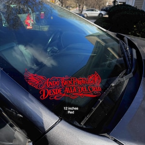 May include: A red vinyl decal with the text "Ando Bien Protegido Desde Alla Del Cielo" and a wing design is displayed on a car windshield. The decal measures 12 inches. The car is dark gray.