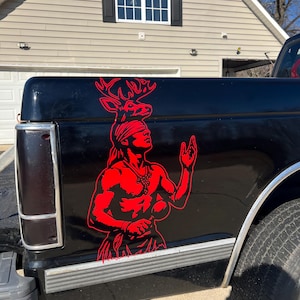May include: A black truck bed features a red graphic of a Native American figure with a deer head. The figure is depicted with a headdress and necklace, arms raised. The truck has a silver trim and a black bumper.
