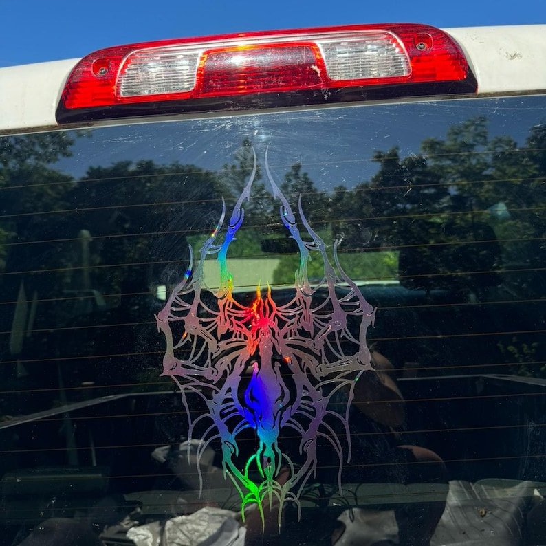 May include: A holographic car sticker with an elaborate spiderweb design. The sticker has a central, flame-like shape with rainbow colour gradients. The sticker is attached to the rear window of a vehicle, with the brake light visible above.