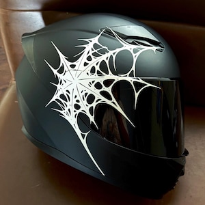 Puede incluir: Un casco de moto integral en color negro mate con un gráfico blanco prominente de una telaraña estilizada que se extiende por el lateral y la parte superior. El casco incluye una visera tintada oscura y un diseño de carcasa liso y aerodinámico.