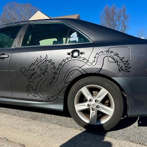 Puede incluir: Un coche gris con una gran calcomanía de dragón negro en el lateral. El diseño detallado del dragón cubre una parte significativa del panel lateral del vehículo, extendiéndose desde la puerta delantera hasta la trasera.