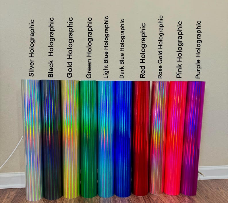 May include: A selection of holographic vinyl rolls in a range of colours: silver, black, gold, green, light blue, dark blue, red, rose gold, pink, and purple. Each roll is labelled with its colour name.