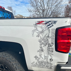 Könnte beinhalten: Ein weißer Pickup mit einer schwarz-weißen Grafik einer Maya-Figur an der Seite. Die Grafik ist detailliert und enthält komplizierte Designs. Der LKW hat einen schwarzen Reifen und eine schwarze Felge.