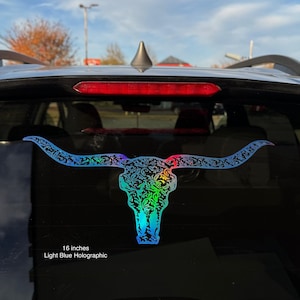 Puede incluir: Una pegatina holográfica azul claro de una calavera de cuernos largos con intrincados patrones florales. La pegatina mide 40.6 cm y está adherida a la ventana de un coche. Efecto arcoíris.