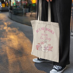 May include: Cream-coloured tote bag with pink text reading "BUY ME BOOKS & TELL ME I'M PRETTY". The bag displays a cartoon bunny reading a book, with books and a cake illustrated around it.