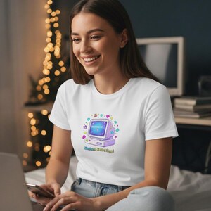 Puede incluir: Camiseta blanca con un gráfico de ordenador retro, con el texto "Waiting to Boot Up!" y "Status: Rebooting!". El gráfico incluye corazones, estrellas y un ordenador morado. La persona sonríe mientras usa un portátil.