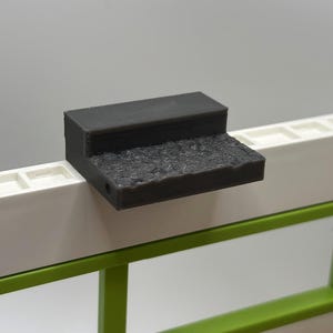Puede incluir: Un escalón de plástico gris oscuro con una superficie texturizada. El escalón está unido a una superficie blanca con un borde verde.