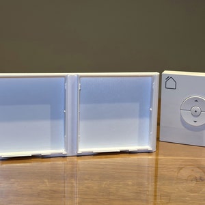 May include: A white Velux window blind remote control with a round button and a house icon. The remote is next to a white plastic cover with two square openings.