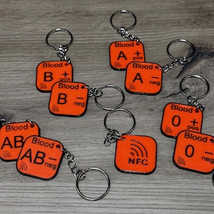 May include: A collection of orange keychains with black text indicating blood types, including A+, A-, B+, B-, AB+, AB-, and O+ and O-. Each keychain has a silver ring and chain.