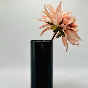 Könnte beinhalten: Eine schwarze zylindrische Vase mit einer einzigen rosa Blume.