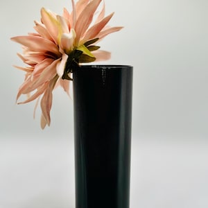 Puede incluir: Una sola flor rosa en un jarrón cilíndrico negro. La flor está parcialmente fuera del jarrón y se inclina hacia un lado.