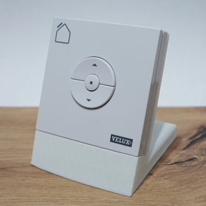 Può includere: Comando per finestra VELUX bianco con un pulsante rotondo e un'icona di una casa. Il comando è su un supporto bianco.
