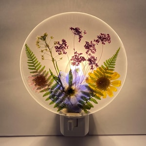 Peut inclure: Veilleuse ronde avec un design transparent orné de fleurs séchées. Les fleurs comprennent des fleurs violettes, jaunes et blanches, ainsi que des feuilles de fougère vertes. La veilleuse a une base blanche et une prise.