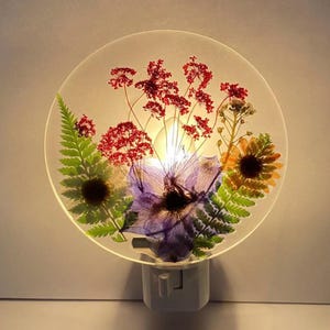Peut inclure: Veilleuse décorative circulaire avec des fleurs pressées. L'arrangement comprend des fleurs rouges, violettes et jaunes, ainsi que des fougères vertes, sur un fond translucide. La veilleuse a une prise blanche.