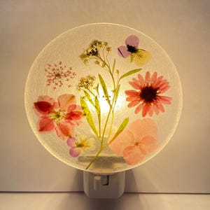 May include: A round night light with a white base, featuring a translucent face embedded with dried flowers in shades of pink, red, yellow, and purple. The light shines through the flowers, creating a soft glow.