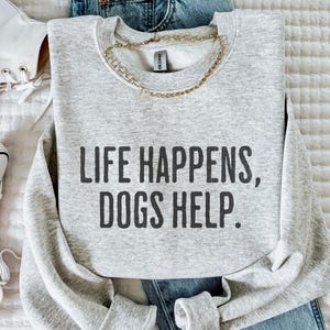 May include: A light gray sweatshirt with the text "LIFE HAPPENS, DOGS HELP." is displayed with a pair of white high-top sneakers and blue jeans. A gold chain necklace is draped on the sweatshirt.