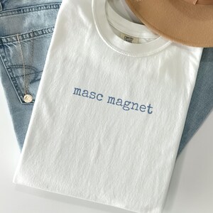 May include: A white t-shirt with the text "masc magnet" in blue. The shirt is folded and placed on top of blue jeans, with a gold necklace and a tan hat in the background. The shirt is made of a soft material.
