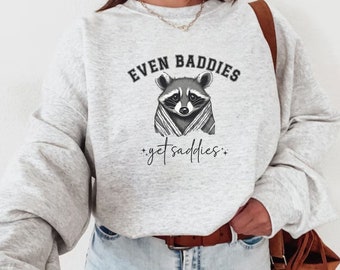 May include: A gray sweatshirt with a raccoon wearing a blanket and the text "Even Baddies Get Saddies".