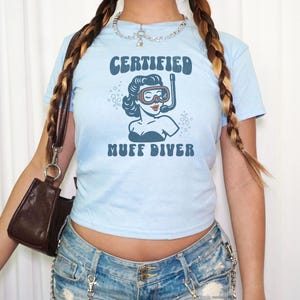 May include: Light blue t-shirt with the words "CERTIFIED MUFF DIVER" and a graphic of a woman wearing a diving mask and snorkel. The shirt is short-sleeved and has a round neckline. The image is of a person wearing the shirt.