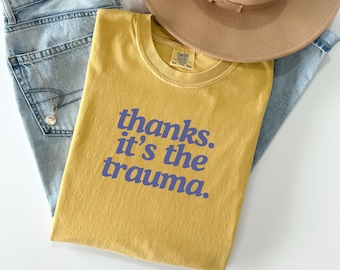 Può includere: Una maglietta gialla con la scritta "thanks. it's the trauma." stampata in viola sul davanti.