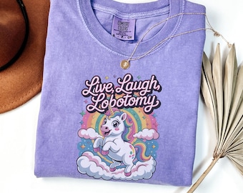 May include: A violet Comfort Colors t-shirt featuring a unicorn graphic with a rainbow and the text "Live, Laugh, Lobotomy." The shirt has a crew neck and short sleeves. A gold necklace is draped across the shirt.
