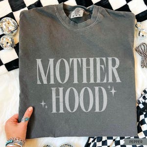 May include: Pepper-colored t-shirt with the word "MOTHERHOOD" in a light grey font. The shirt has a crew neck and short sleeves. The text is accompanied by starburst graphics. The shirt is displayed on a white fur-like surface.