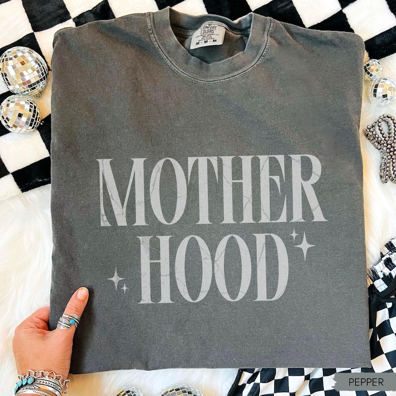 May include: Pepper-colored t-shirt with the word "MOTHERHOOD" in a light grey font. The shirt has a crew neck and short sleeves. The text is accompanied by starburst graphics. The shirt is displayed on a white fur-like surface.
