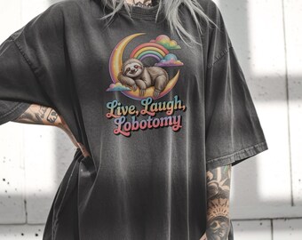 May include: Dark gray t-shirt featuring a sloth resting on a crescent moon with a rainbow and clouds. The shirt has the text "Live, Laugh, Lobotomy" in a colorful, retro font. The shirt is a relaxed fit.