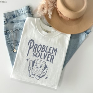 May include: White t-shirt featuring the words "PROBLEM SOLVER" and a cartoon calculator graphic. The shirt is styled with light blue jeans and a beige hat. The graphic includes a ruler and pencil.