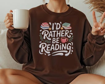May include: Dark chocolate brown sweatshirt with the text "I'd Rather Be Reading" in white script. The design includes stacks of books, a coffee cup, and decorative leaves. A white mug is held in the left hand.