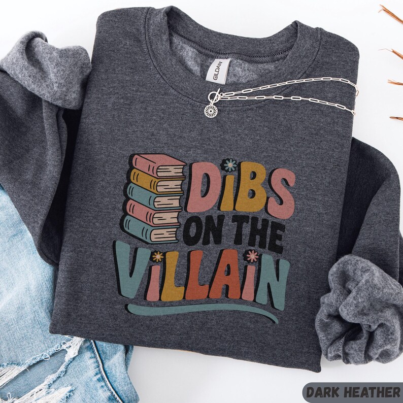 May include: Dark heather gray sweatshirt with the text "Dibs on the Villain" in colorful retro lettering. The design includes a stack of books. A silver necklace with a sun pendant is draped across the sweatshirt.