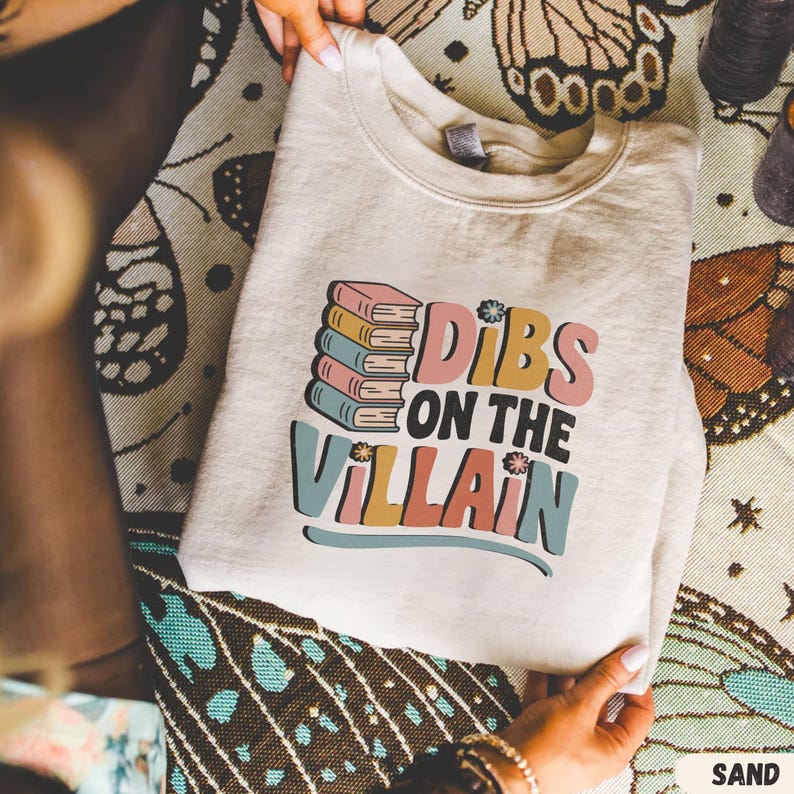 May include: Sand-colored sweatshirt with the text "Dibs on the Villain" in a colorful, retro font. The design includes a stack of books in pink, blue, and yellow. The sweatshirt is folded and held by hands.