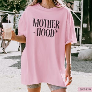May include: A light pink oversized t-shirt with the words "MOTHER + HOOD" printed in black. The shirt has a relaxed fit and short sleeves. The text is accompanied by star symbols. The shirt is made of a soft material.
