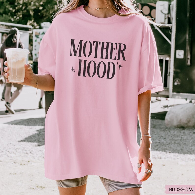 May include: A light pink oversized t-shirt with the words "MOTHER + HOOD" printed in black. The shirt has a relaxed fit and short sleeves. The text is accompanied by star symbols. The shirt is made of a soft material.