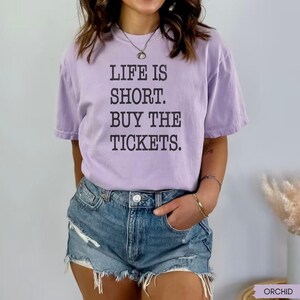 Puede incluir: Una camiseta lila con el texto negro "LIFE IS SHORT. BUY THE TICKETS.". La camiseta se combina con pantalones cortos de mezclilla. La imagen presenta un estilo casual y relajado.