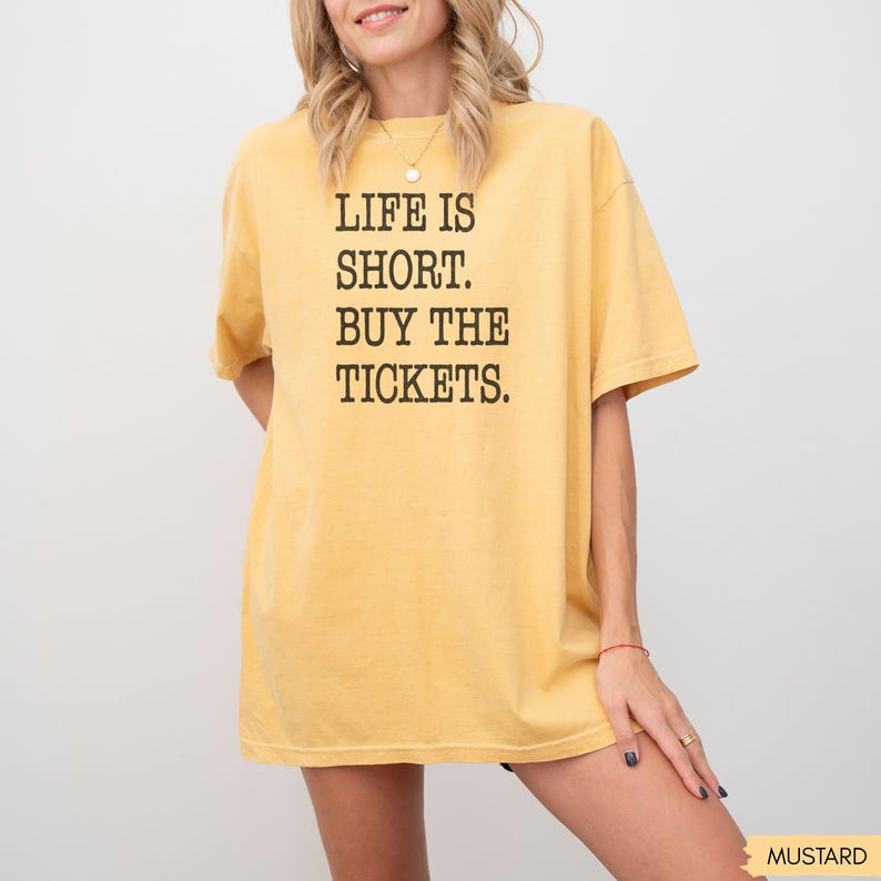 Puede incluir: Camiseta amarilla mostaza con el texto "LIFE IS SHORT. BUY THE TICKETS." en letras negras. La camiseta es de estilo casual y de gran tama&ntilde;o.